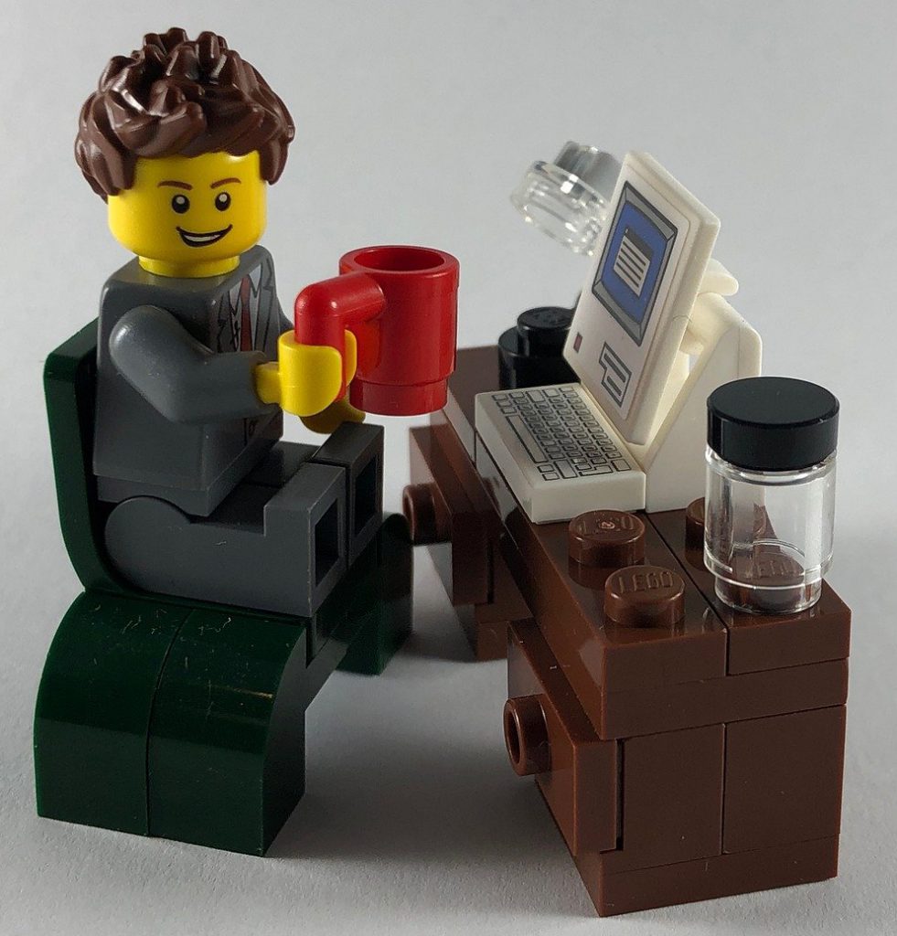 This is a picture of a Lego businessman sat at a brown desk with a red cup of coffee in his right hand, computer on the desk, a desk lamp and a jar of biscuits.